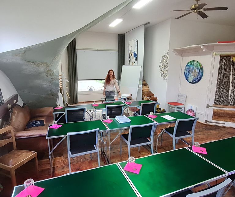 Frau mit langen roten Haaren steht in einem hellen Raum mit mehreren grünen Tischen, schwarzen Stühlen und einem Flipchart neben einem Fenster unter einer Dachschräge.